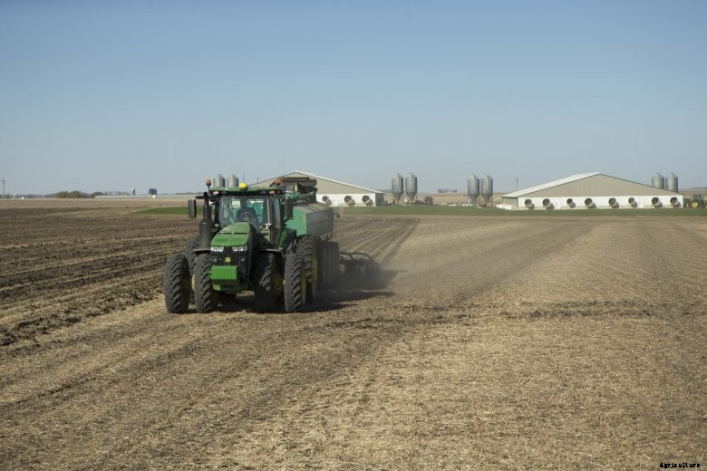 A John Deere tractor pulls a liquid manure tank over a field with hog confinements in the background